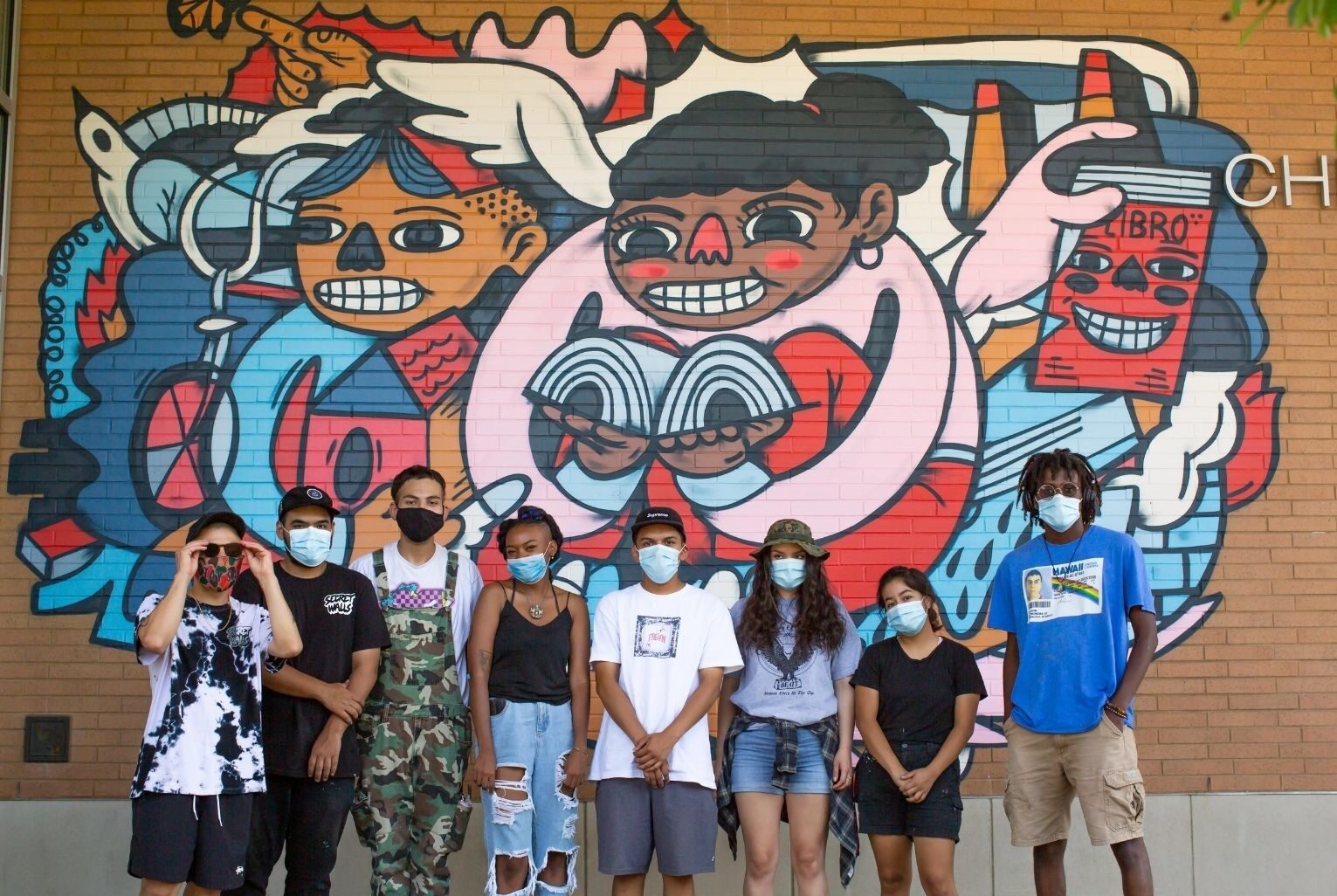a group of young artists of varied ages with masks on standing in front of a colorful mural