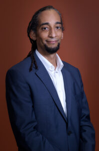 A formal portrait of Asif Wilson in a suit, against a maroon backdrop