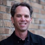 Portrait of Brian Dolinar in a dark shirt standing next to a brick wall