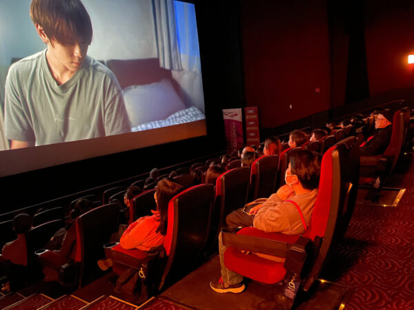 A theatre full of movie goers watch a film at a screening hosted by Asian Pop Up Cinema in Chicago. On screen a character sits in their home, looking to the distance.