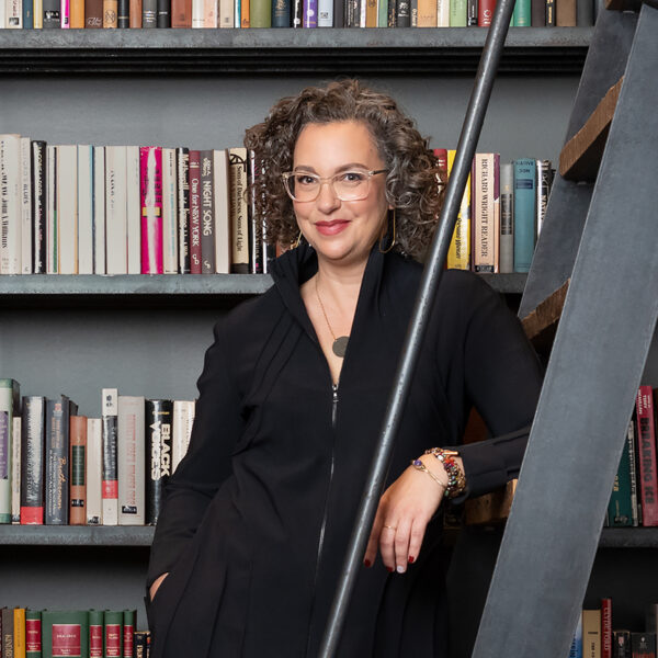 Executive Director Gabrielle Lyn poses on a ladder in a library, a wall of books behind her