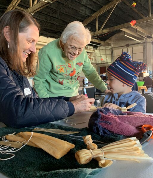 Adults and child gathered around a table making corn husk dolls